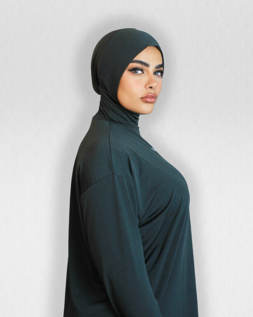 Model wearing RUUQ Oversized Long sleeve Top with high neck Hijab Cap attached in Forest Green. Thumb holes at cuff. Curved scoop hem covers past mid thigh. RUUQ logo at front center and RUUQ emblem at back center.
